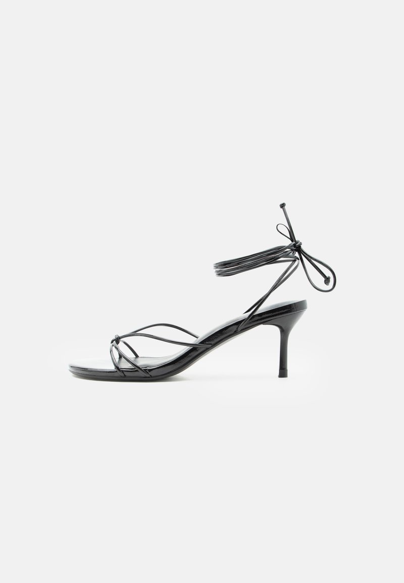 Μαύρο σανδάλι με λεπτά λουράκια, ψηλό τακούνι στιλέτο μεσαίου ύψους και λεπτούς δεσίμους στον αστράγαλο σε λευκό φόντο.