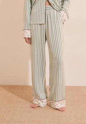 Una persona indossa pantaloni del pigiama a righe verdi e bianche con polsini floreali, in piedi a piedi nudi su un pavimento beige strutturato.