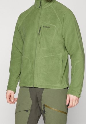 Mies, jolla on päällään vaaleanvihreä Columbia-villatakki ja oliivinvihreät cargo-housut, yksiväristä taustaa vasten.