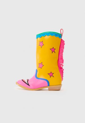 Knæhøye cowboy støvler i gul med rosa aksenter, med stjernesymboler, bølget topp, frynser og et smilende design på tåen.