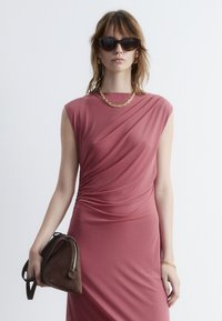 Rosafarbene, drapierte Kleid mit kurzen Ärmeln, glattem Stoff und gerafften Details. Kombiniert mit einer braunen Wildledertasche in Triangel-Form und Sonnenbrille.