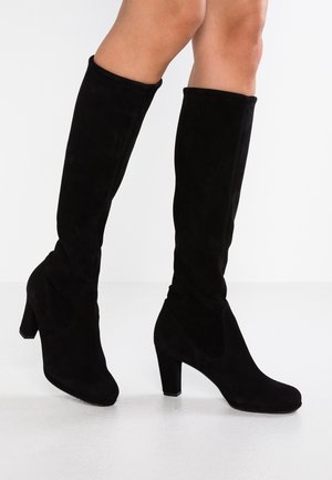 Femme portant des bottes hautes en daim noir à talons, debout sur un sol blanc, jambes croisées.
