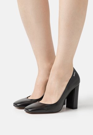Czarne skórzane buty na wysokim obcasie z kwadratowym noskiem i masywnym obcasem. Subtelne logo na obcasie oraz gładka, wypolerowana faktura.