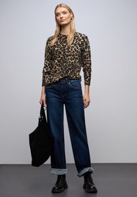 Langärmeliges Oberteil mit Leopardenmuster in Beige und Schwarz; kombiniert mit dunkelblauen Jeans mit weitem Bein, an den Säumen umgeschlagen, und schwarzen Schnürstiefeln. Schwarze Tasche.
