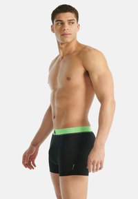 Pánské boxery v černé barvě s zeleným elastickým pasem, které mají hladkou texturu a přiléhavý střih. Pas má jemný vzor.