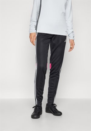 Personne portant un pantalon de survêtement Adidas noir avec des bandes blanches et un haut blanc à manches longues, debout sur un fond clair uni.
