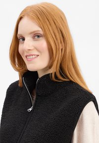 Zwarte fleece bodywarmer met hoge kraag, ritsluiting en een gestructureerd oppervlak. Draagt over een lichtgekleurde top, wat een modern ontwerp toont.