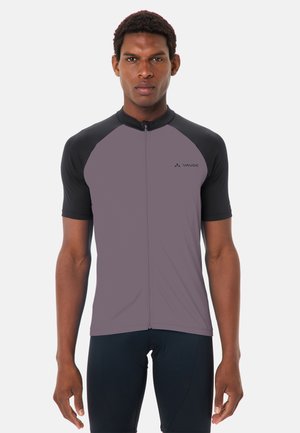 Uomo in piedi, rivolto verso avanti, che indossa una maglia da ciclismo a maniche corte viola e nera con una cerniera frontale e pantaloncini da ciclismo neri.