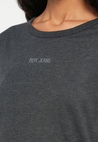 Közelkép egy női dekoltázsról, aki sötétszürke, kerek nyakú pólót visel, a mellkason középen kicsi, fehér felirattal: "PEPE JEANS".