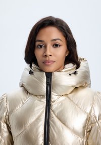 Metallic guld pufferjakke med høj krave, vandret quiltet mønster og sort lynlås lukning. Glat, skinnende tekstur med snore.