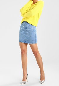 Blaue Jeans-Minirock mit Sternenverzierungen, kombiniert mit einem leuchtend gelben Strickpullover. Silberne hohe Absätze vervollständigen das Outfit.