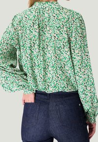 Blusa floral con estampado de leopardo en verde y beige, mangas largas, escote fruncido y corte relajado, combinada con jeans oscuros de mezclilla.