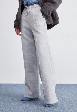 Person trägt lockere, hellgraue Jogginghosen mit Kordelzug und blaue Schuhe mit Absatz, steht auf einem blau teppichboden vor einer weißen Wand.