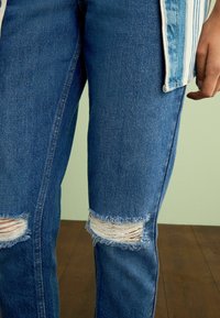 Rebitud sinine denim teksad, millel on frayed augud põlvede juures, sirge jalgade lõikega ja kerge narmenduse tõttu, mis lisab tekstuuri.
