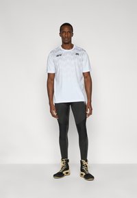 Λευκό T-shirt για προπόνηση με γεωμετρικό σχέδιο, μαύρο λογότυπο UFC. Μαύρο κολάν με ποικίλες υφές, συνοδευόμενο από μαύρα και χρυσά μποτάκια ψηλού πάτου.