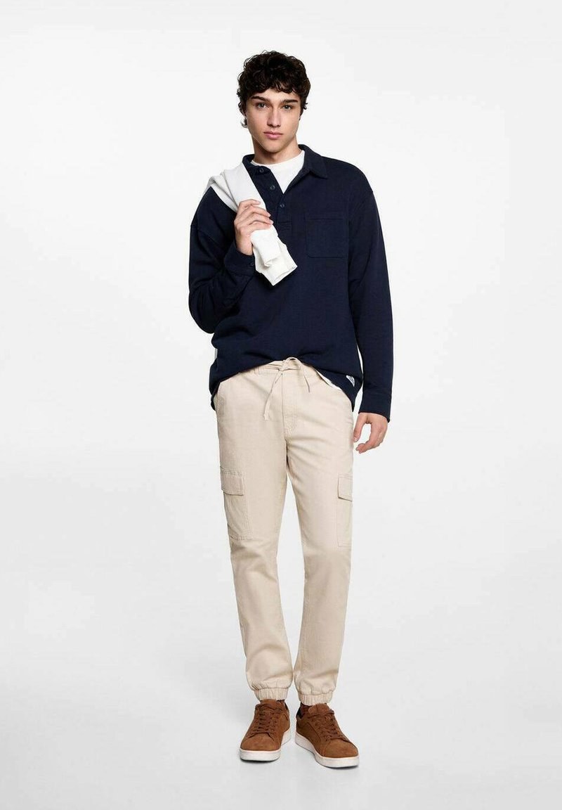 Mörkblå långärmad polo, beige cargobyxor med elastiska muddar och bruna sneakers. Modell håller en vit sweatshirt, neutral bakgrund.