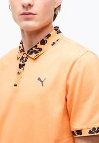 Homme portant un polo pêche avec un motif floral noir sur le col et les poignets des manches, arborant un petit logo Puma sur la poitrine.