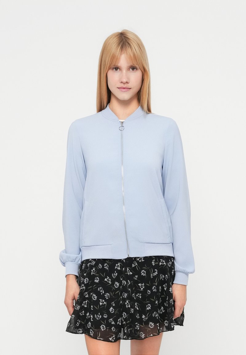 Veste bomber bleu clair avec fermeture éclair avant, poignets côtelés et poches latérales, associée à une jupe noire à motif floral avec une coupe évasée.