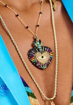 Goldene Halskette mit einem herzförmigen Anhänger. Der Anhänger ist mit bunten Perlen, einem Augendesign und einem strukturierten schwarzen Hintergrund versehen.