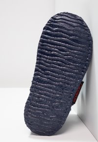 Zool van een slipper, zwart met een gestructureerd golvend patroon. Heeft een rood label aan de zijkant. Materiaal lijkt rubberachtig met groeven voor grip.