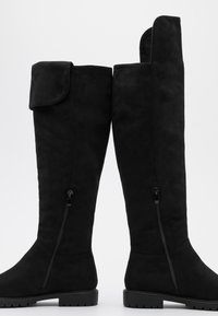 Botas de ante negras hasta la rodilla con suela plana, cremalleras laterales y parte superior doblada. Textura suave sin patrones o acentos visibles.