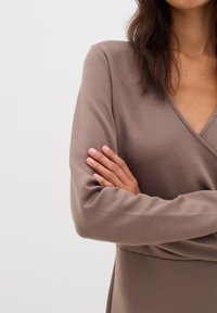Hnědé dlouhé šaty s dlouhým rukávem, výstřihem do V a zavinovacím designem, vyrobené z hladkého, pružného materiálu. Ruce jsou zkřížené vpředu.