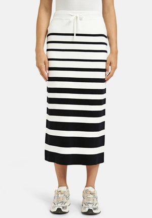 Frau trägt einen weißen Midirock mit schwarzen horizontalen Streifen und weißen Turnschuhen, steht mit entspannten Händen an den Seiten.