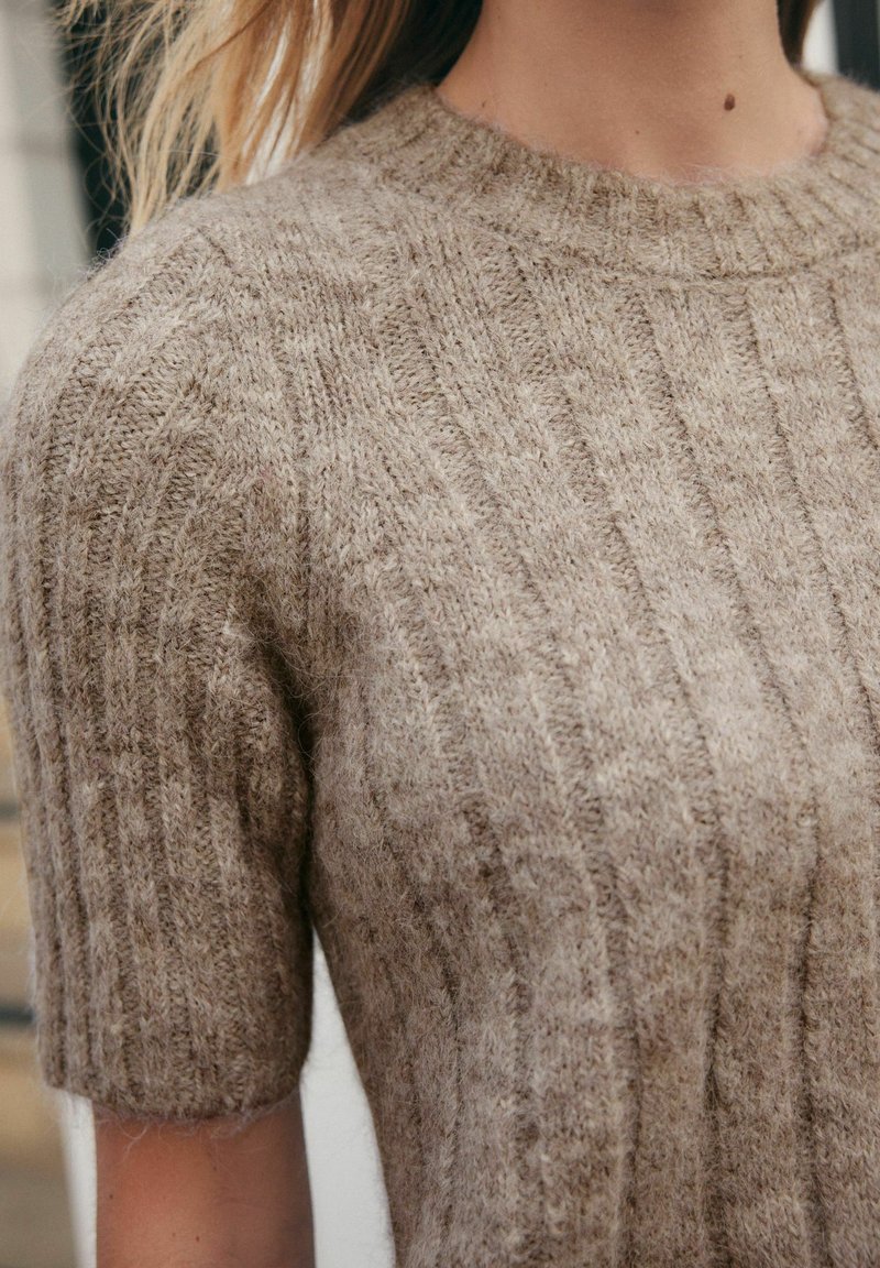 Maglione lavorato a maglia color marrone chiaro, con texture a costine e maniche corte. Il tessuto sembra morbido e leggermente peloso con uno spessore medio.