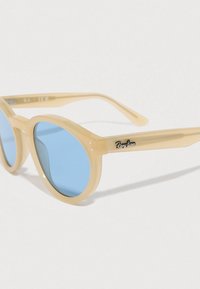 Kerek napszemüveg világos bézs műanyag kerettel és kék lencsékkel, oldalán fekete Ray-Ban logóval, sima felületű.