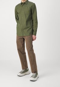 Camisa verde de manga larga con cierre de botones y bolsillo, combinada con pantalones marrones y zapatillas verdes con suelas blancas y cordones negros.