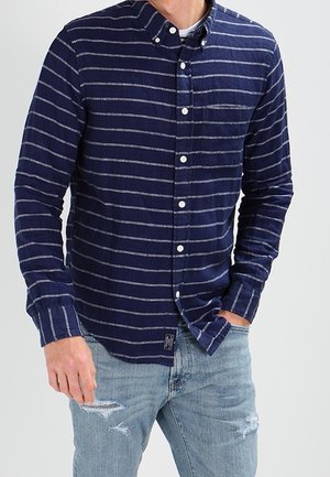 Mann trägt ein marineblaues Hemd mit weißen horizontalen Streifen und hellblauen, zerrissenen Jeans, steht mit einer Hand in der Tasche.