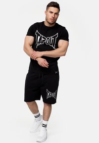 Juodas medvilninis marškinėlių ir šortų komplektas su baltu "TAPOUT" logotipu. Savybės: apvalus kaklo iškirpimas, elastinga juosta ir šoninės kišenės. Baltos sportbačiai ir dryžuotos kojinės.