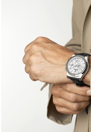 Hombre ajustando un reloj de pulsera cronógrafo plateado con una correa negra, vistiendo una chaqueta beige contra un fondo claro y liso.