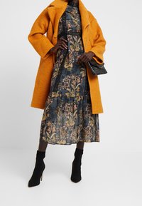 Senfgelber Wollmantel über einem Kleid mit dunklem Blumenmuster und Spitzenakzenten, kombiniert mit schwarzen Knöchelschuhen und einer kleinen strukturierten schwarzen Handtasche.