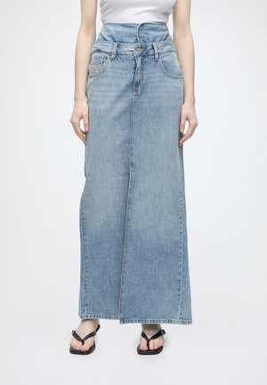 Femme portant une jupe longue en denim bleu clair taille haute avec double ceinture, associée à des sandales noires et un haut blanc.