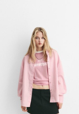 Camicia a righe rosa oversize con bottoni sopra una t-shirt grafica rosa e una gonna nera. Tessuto morbido con vestibilità rilassata e design informale.