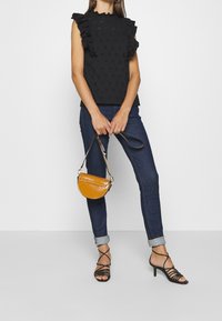 Svart ärmlös topp med volanger vid axlarna och blommiga hålmönster, i kombination med mörka jeans och en liten glansig orange handväska.