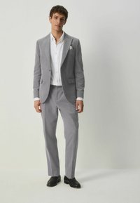 Traje de pana gris claro con solapa muesca, cierre de botón y bolsillos laterales, combinado con una camisa blanca de botones y zapatos negros.