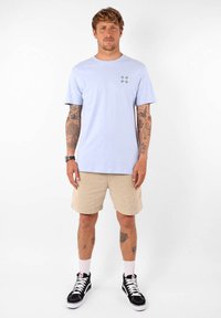 T-shirt en coton bleu clair à manches courtes, avec un petit motif sur la poitrine gauche. Associé à un short beige et des baskets noires.