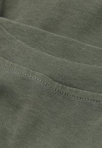 Zelená bavlněná tkanina s hladkou strukturou. Má lemovaný okraj a jemný šev, přičemž zobrazuje detailní pohled na materiál.