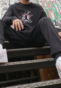 Schwarzes Langarmshirt mit buntem Grafikdesign, kombiniert mit schwarzen Hosen und weißen Sneakern mit pinken Akzenten, sitzt auf Metallstufen.