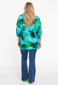 Losse, kleurrijke top met blauwe en groene abstracte patronen, gecombineerd met uitlopende denim jeans en sandalen met groene accenten.