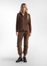 Hnědá fleecová bunda na zip s odpovídajícími kalhotami, která má měkkou texturu, elastické rukávy a dvě boční kapsy, spárovaná s černými teniskami.