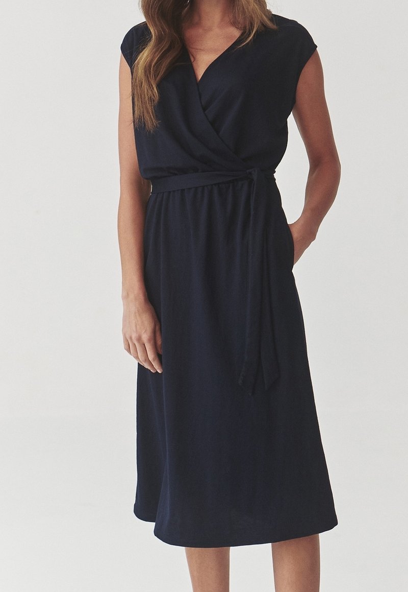 Femme portant une robe portefeuille noire sans manches avec un col en V et une ceinture nouée à la taille, debout avec une main dans la poche sur un fond uni.