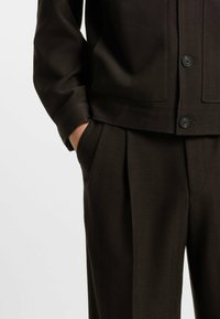 Dark brown herringbone suit jacket with button closure, side pockets, and matching trousers featuring pleats and a tailored fit.