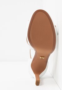 Vue de la semelle d'une chaussure à talon blanc avec une semelle marron et un petit logo doré près de la voûte plantaire, sur fond blanc.