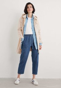 Manteau beige, chemise en denim bleu clair, débardeur blanc, jean bleu ample et baskets blanches avec des accents violets. Tenue décontractée en couches.