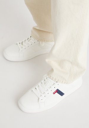 Zapatillas sintéticas blancas con suela plana, que presentan una franja de acento roja y azul marino y detalles de superficie texturizada. Combinadas con pantalones beige claros.