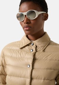Beżowa pikowana kurtka z kołnierzem i metalowymi zatrzaskami, zestawiona z białymi owalnymi okularami przeciwsłonecznymi z lustrzanymi soczewkami.