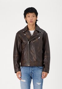 Giacca motociclista in pelle marrone con zip asimmetrica, colletto a scatto e tasca frontale sul petto. Indossata sopra una maglietta bianca e jeans blu.